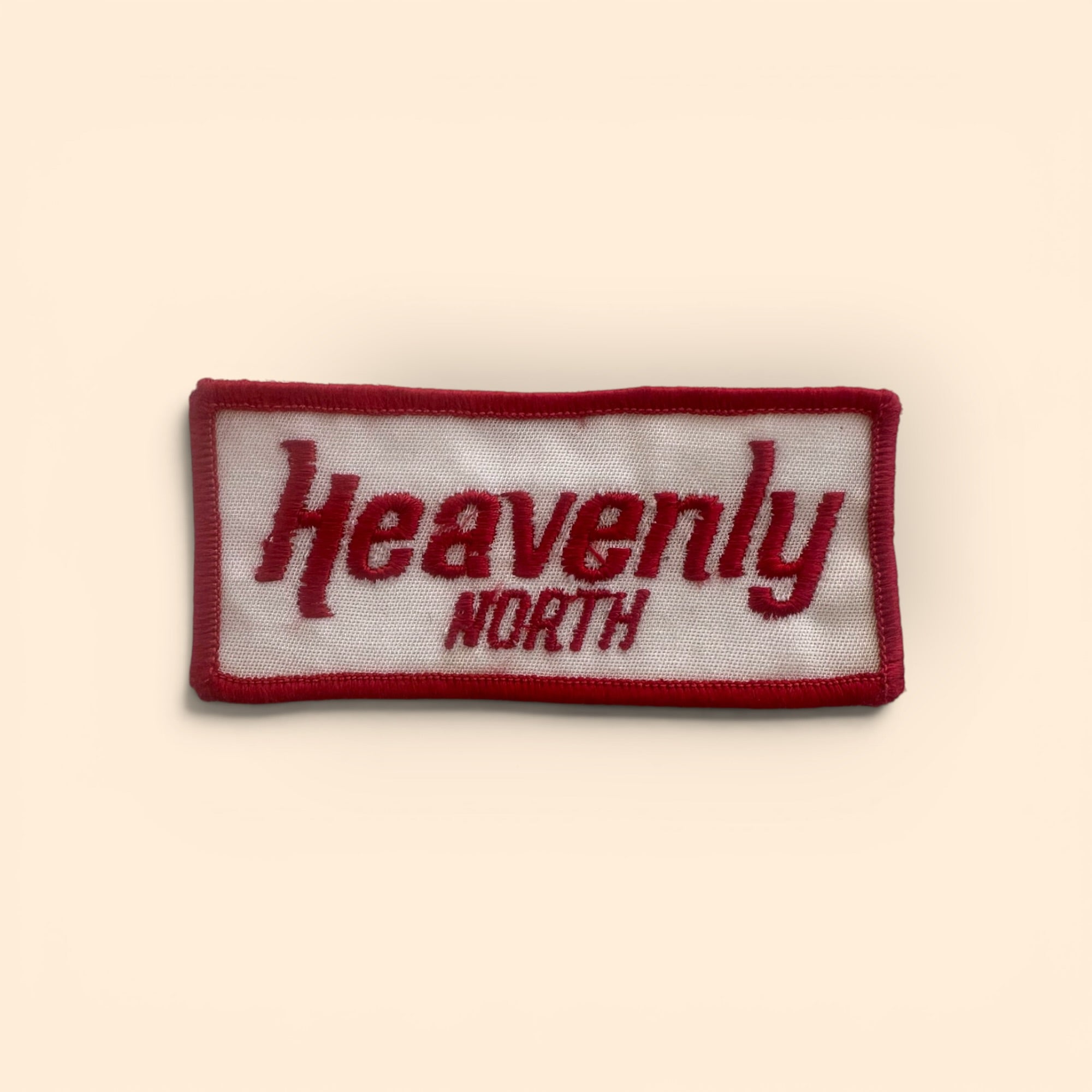 A rectangular embroidered patch with a white background and a dark red border, featuring the words 'Heavenly NORTH' in dark red embroidery, shown on a light beige background.