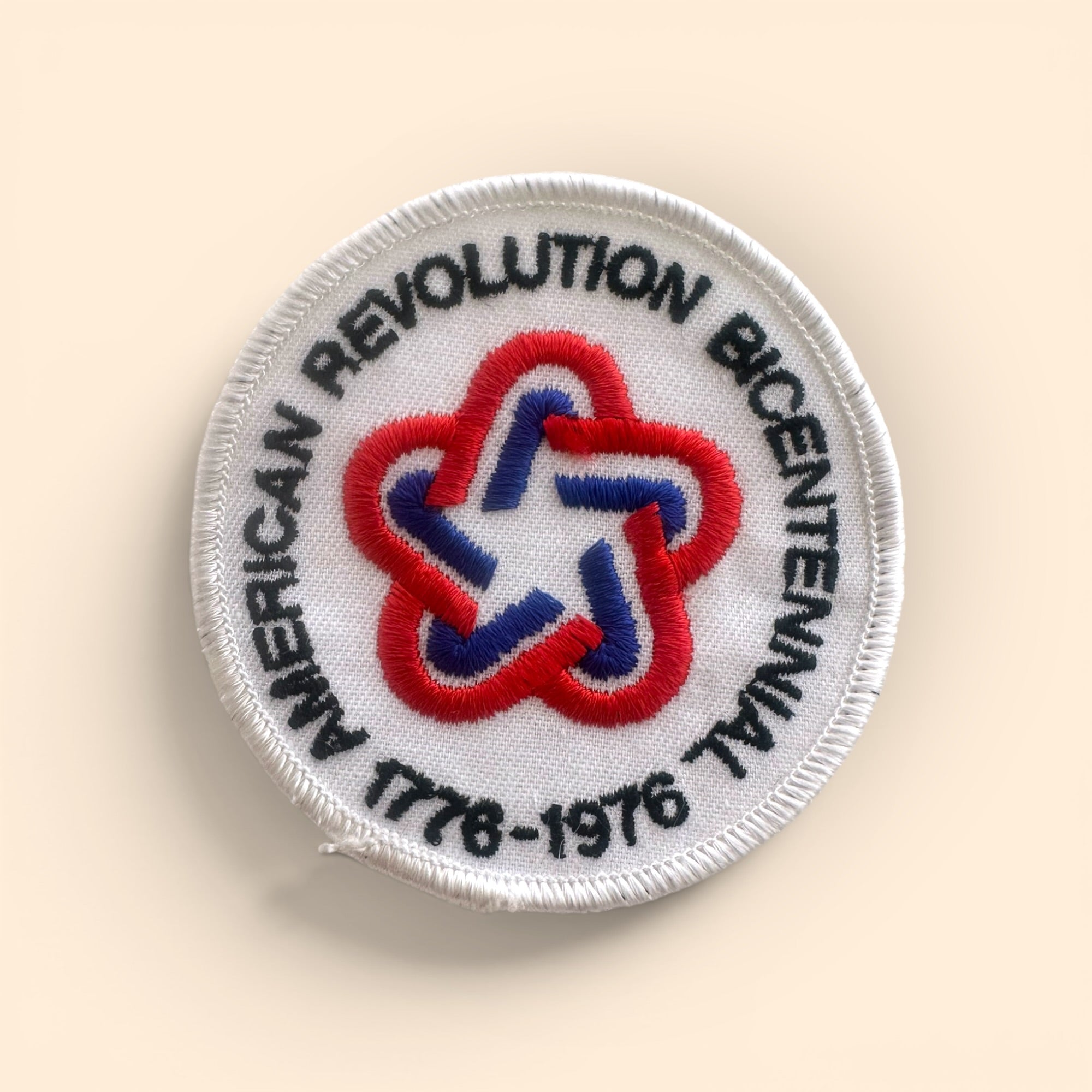 A circular embroidered patch with a white border and text reading 'AMERICAN REVOLUTION BICENTENNIAL 1778-1976' around a red, white, and blue star motif, shown on a light beige background.