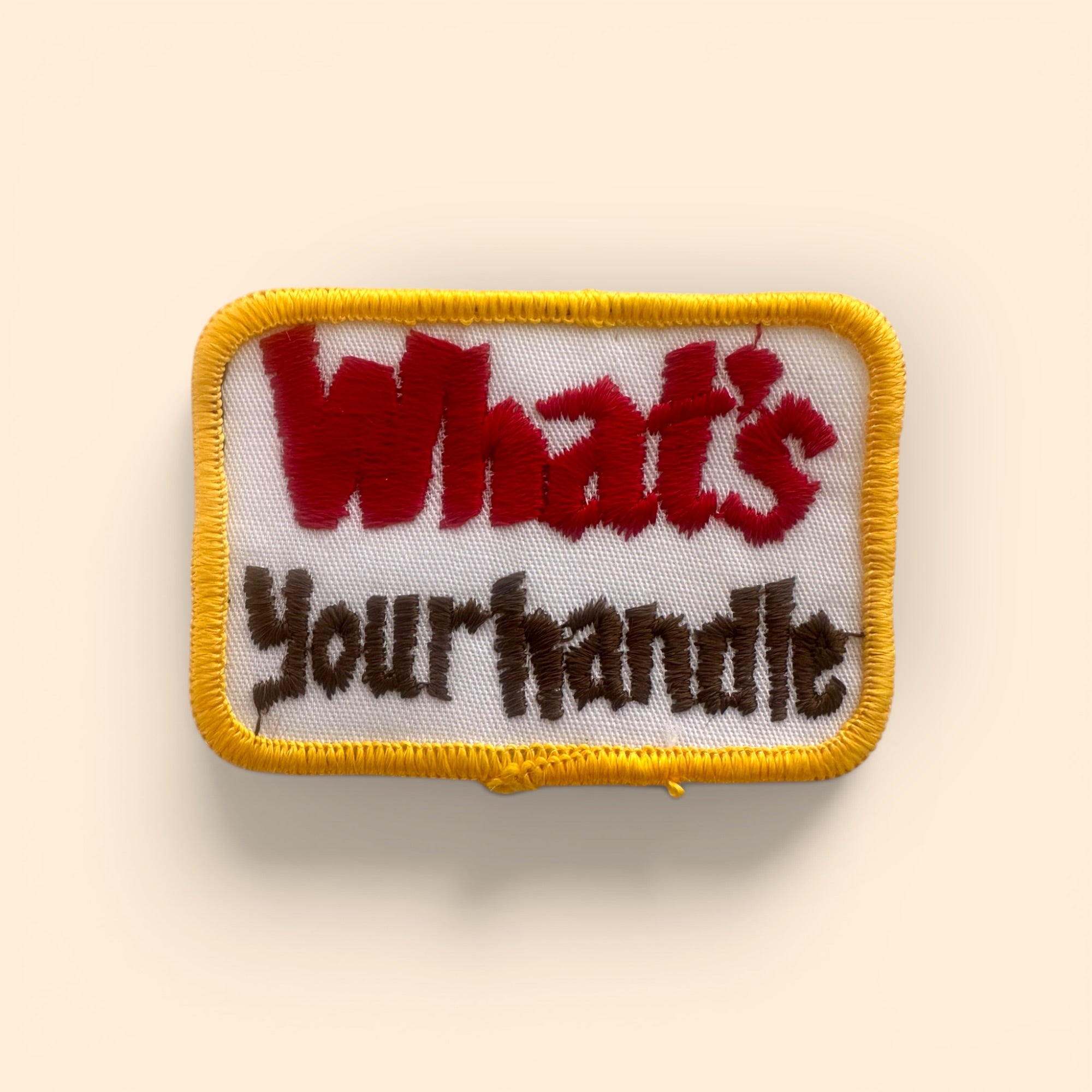 An embroidered rectangular patch with a yellow border, featuring the text 'What's Your handle' in red and dark brown lettering on a white background, set against a light beige backdrop.